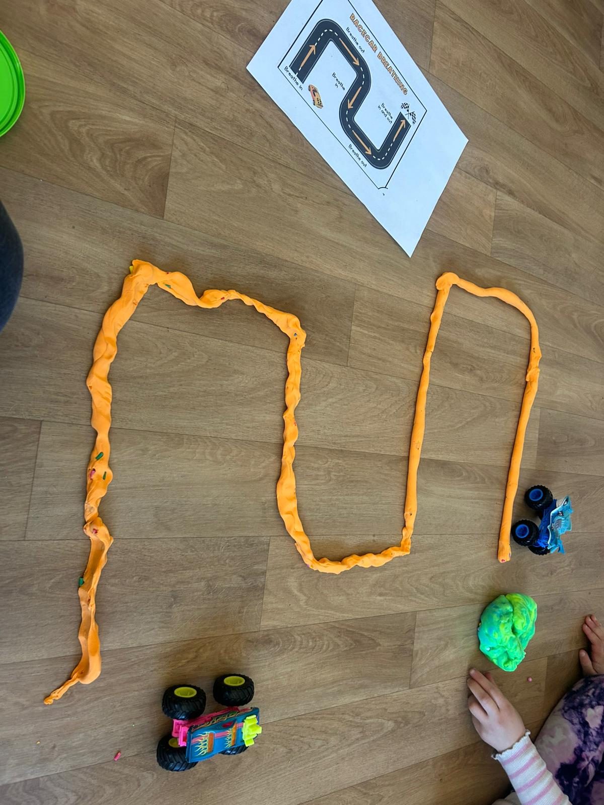 A long orange playdough racetrack laid out on the floor next to a printed racecar breathing sheet, with toy cars and green playdough beside a child's hands