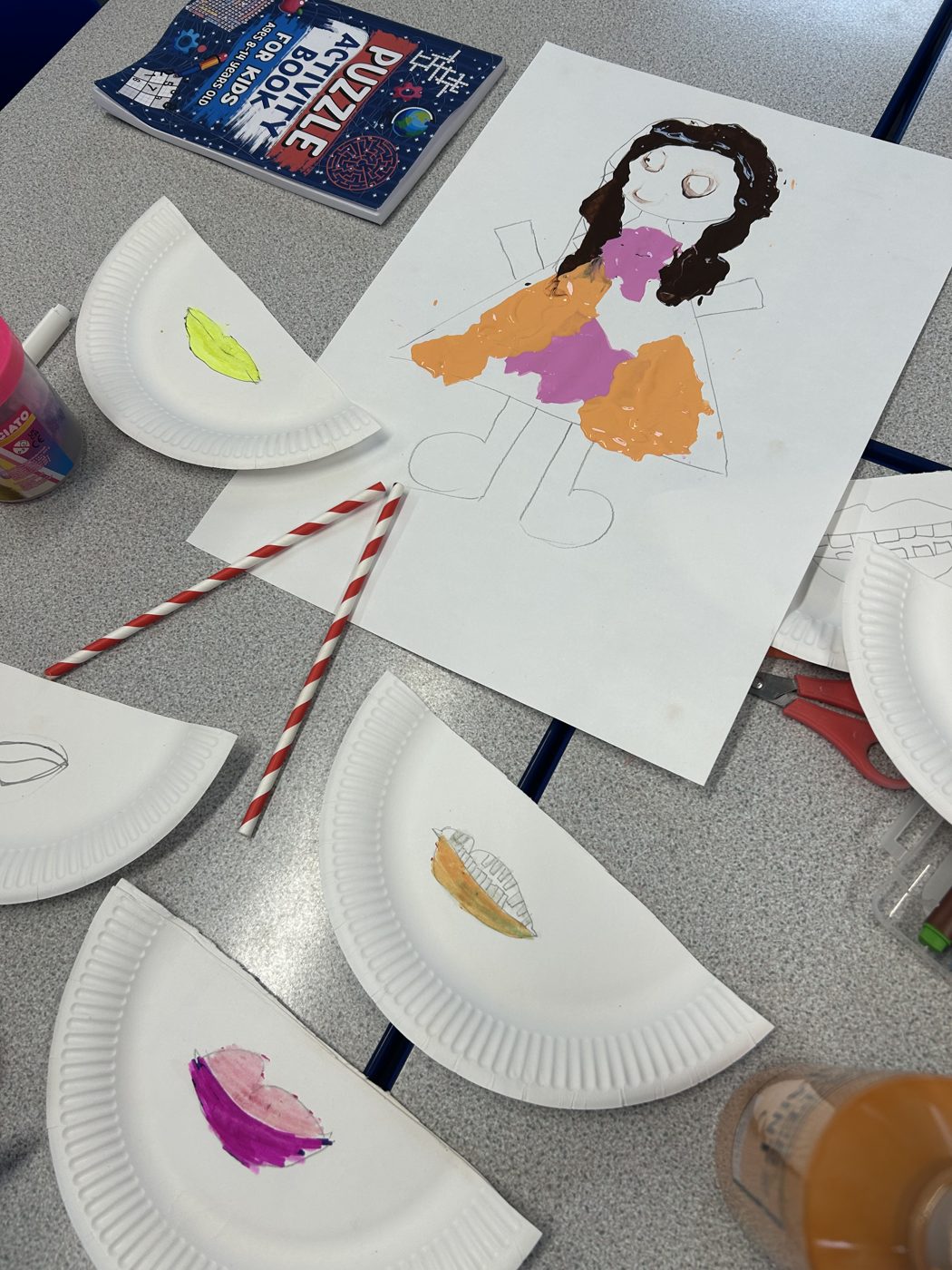 A table set up for a coaching activity with paper plates, coloured paints, straws and a child's painted self portrait