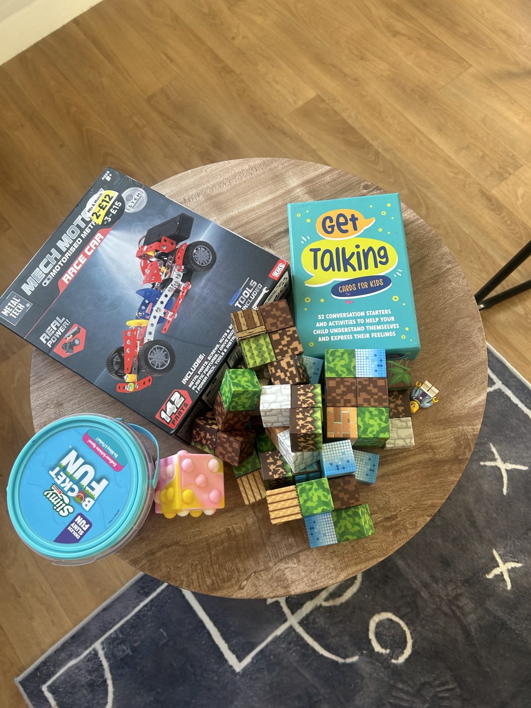 A round wooden table with building blocks, a motorised race car model kit, slime and Get Talking conversation cards for children